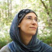 Headshot of woman in glasses wearing a hijab. Trees and bushes in the background