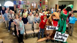 Multiple people stand in a room looking towards the camera, with two people in dinosaur suits at the front. The dinosaurs are holding signs that say "Today" and "Evolution Sunday."