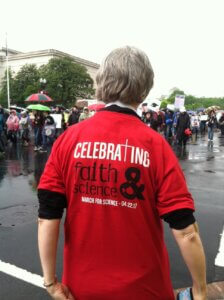 A woman standing with her back to the camera, wearing a red shirt that says "Celebrating Faith & Science." There are people on a street in the background holding umbrellas.